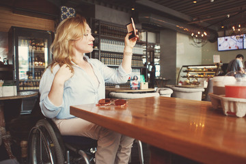 Woman in a wheelchair is drinking coffee and making selfie on a smartphone in a cafe.