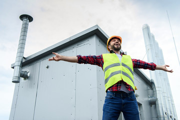 excited male professional constructor in safety vest and helmet posing with open arms on roof