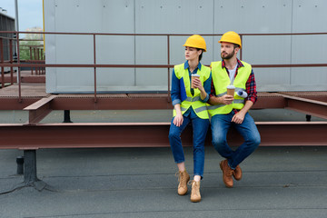 male and female architects in hardhats with blueprint on coffee break sitting on roof