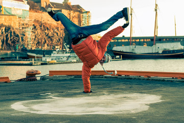 Young guy dancing break dance on the waterfront in an orange jacket in winter.