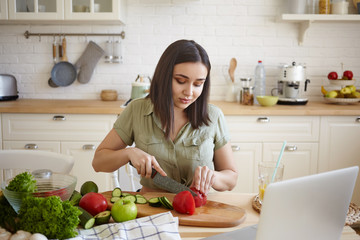 Obraz na płótnie Canvas Portrait of attractive casually dressed young dark haired chubby woman cutting vegetables using sharp and cooking board, sitting at kitchen table, watching video recipe online on laptop computer
