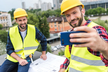 male engineers in safety vests and helmets with blueprints taking selfie on smartphone on roof
