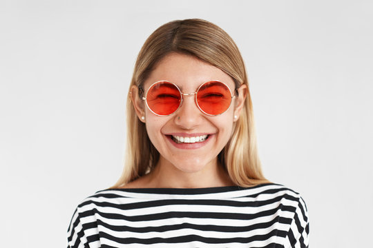 Positiveness, joy and happiness concept. Picture of happy joyful hipster girl in trendy striped top and stylish pink round shaped shades rejoicing at good day or great news, smiling cheerfully