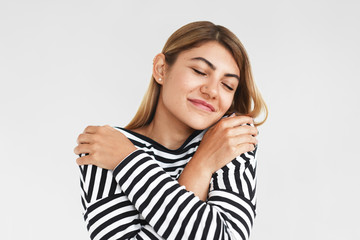 Studio shot of charming positive young woman wearing stylish clothes closing eyes with pleasure, keeping arms around herself, having joyful facial expression, standing isolated at blank wall