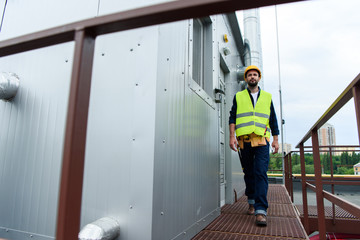 male engineer in safety vest and helmet with tool belt walking on roof