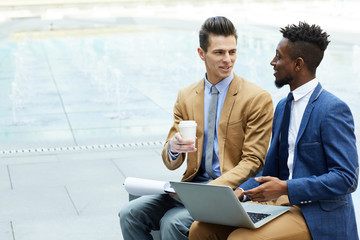 Happy young multiethnic entrepreneurs in stylish outfits discussing business using laptop during...