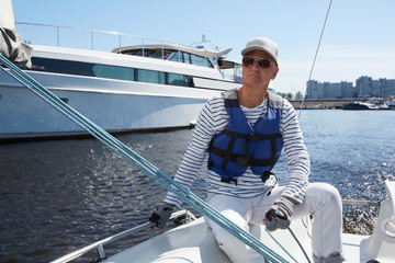 Serious pensive handsome mature male yacht captain in sunglasses and white cap wearing life jacket...