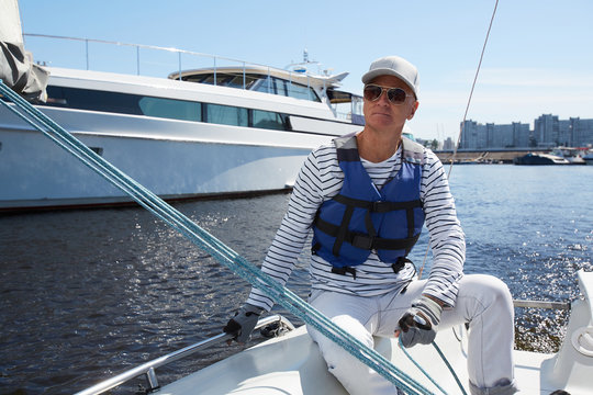 Serious pensive handsome mature male yacht captain in sunglasses and white cap wearing life jacket sitting on boat deck and looking into distance