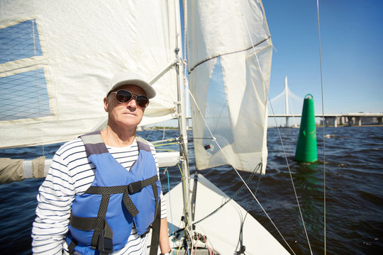 Serious confident experienced mature sailor in sunglasses and cap wearing life jacket looking at camera while standing on yacht