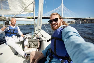 Photo sur Plexiglas Naviguer Cheerful handsome friends in blue life jackets taking selfie against beautiful bridge while sitting on boat deck