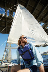 Fototapeta premium Below view of pensive dreamy handsome male sailor in life jacket feeling freedom sitting on sailboat and contemplating waterscape around while passing under bridge