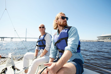 Pensive dreamy male tourists in sunglasses wearing life jackets sitting on sailboat deck and looking around during sailing tour