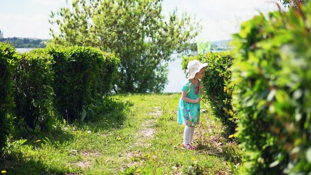 A little sweet girl in a blue dress is catching insects with a green net in the alley next to the river on a sunny summer day.