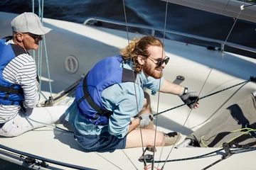 Papier Peint photo Naviguer Happy excited young bearded yachtsman in sunglasses handling sailboat and pulling rope while enjoying sport competition in yacht club