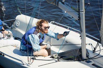Serious focused young bearded sportsman in sunglasses using ropes while directing sailboat during...