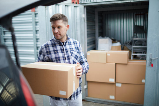 Serious handsome young man in casual clothing finding place in car for cardboard box while loading automobile at container storage area, he storing things in container