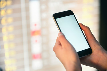 Mockup image of female hands holding black mobile phone with blank white screen over flight board in airport terminal