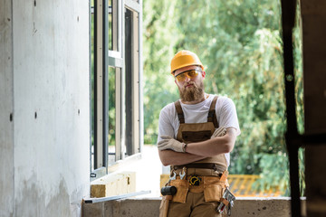 smiling builder in protective googles and hardhat standing with crossed arms at construction site