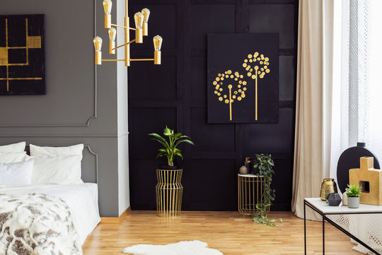 Black and gold poster above plants in bedroom interior with white pillows on bed. Real photo