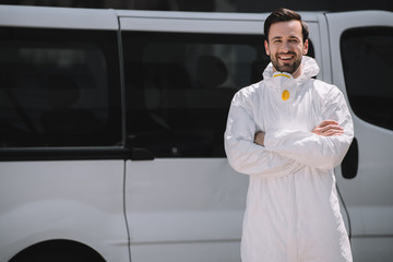 happy pest control worker in uniform standing with crossed arms near car on street