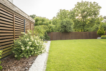 Flowers, trees and green grass in the garden of house with wooden screen. Real photo