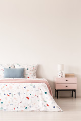 Lamp on cabinet next to patterned bed with blue pillow and pink blanket in bedroom interior. Real photo - obrazy, fototapety, plakaty