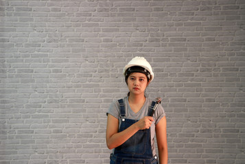 Technician woman ware white helmet with grey T-shirt and denim jeans apron dress standing and steel hammer in hand on grey brick pattern background.