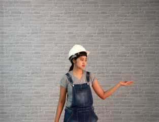 Technician woman ware white helmet with grey T-shirt and denim jeans apron dress standing and palms facing up of left hand on grey brick pattern background.