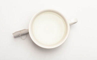 Mug filled with boiling water and teabag on white background. Tea time concept. Cup or white porcelain mug with transparent hot water and bag of tea. Process of tea brewing in ceramic mug, top view