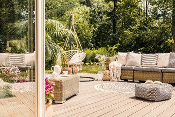 Pouf and rattan sofa on wooden patio with hanging chair in the garden. Real photo - obrazy, fototapety, plakaty