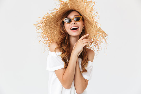 Portrait of modern cute woman 20s wearing big straw hat and sunglasses posing on camera with happy smile, isolated over white background