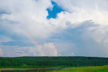 Partial arc rainbow against background of white clouds over green field and forest.