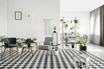 Wooden table between armchairs on checkered floor in white living room interior with plants. Real...