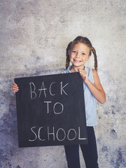 blond schoolgirl is holding blackboard with the words back to school in front of concrete background