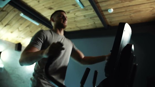 Portrait of male athlete, who is working on his muscules on cross-trainer in the gym. Sportsman in grey t-shirt is moving fast on the equipment in small dark room with wooden ceilings, while making