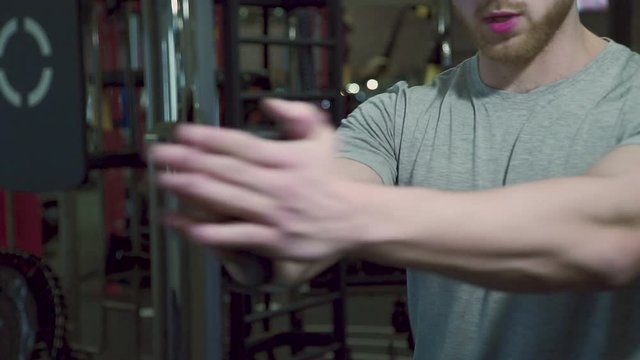 Close up of sportsman, who is doing xable exercise in modern gym. Young male athlete in grey t-shirt is holding handle of weight equipment in his muscular hand and puling it sideway with the turn of