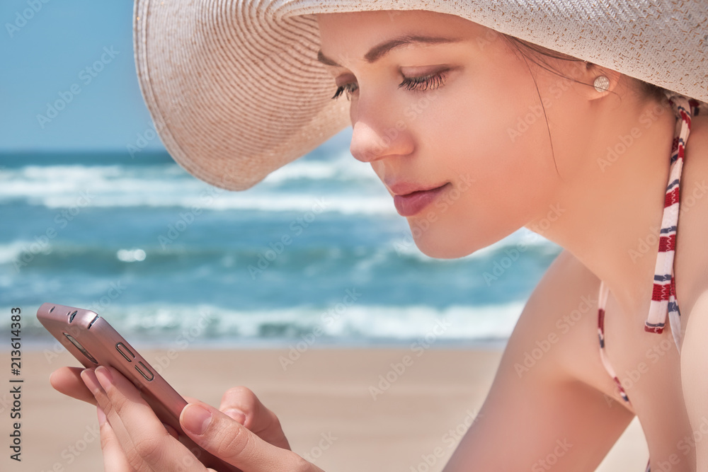 Wall mural beautiful woman in hat using her mobile phone on beach