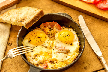 Fried eggs with tomatoes in a cast-iron frying pan and toasted bread on an old wooden table