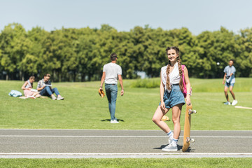 teenage girl with skateboard smiling at camera while friends spending time behind in park