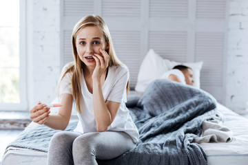 Wonderful news. Alert blond woman holding her pregnancy test and her husband sleeping