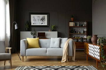 Blanket and pillows on grey settee in retro living room interior with armchair and poster. Real...