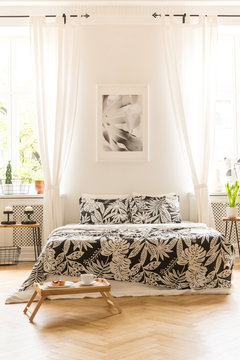 Front view of a double bed with floral sheets, graphic on the wall between windows and breakfast tray on the floor in a hotel room interior