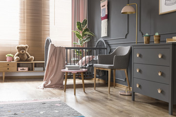 Real photo of a cot standing next to an armchair, lamp and cupboard in dark and classic baby room interior