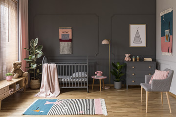Real photo of a grey crib standing next to a pink stool, a lamp and cupboard in grey baby room...