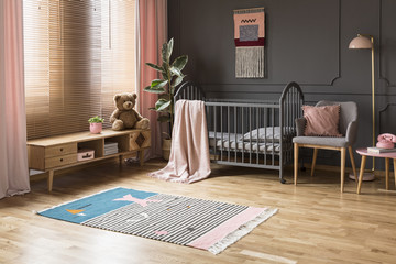 Real photo of a baby crib standing between a low cupboard and an armchair, lamp and stool in...