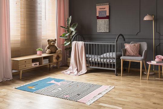 Real photo of a baby crib standing between a low cupboard and an armchair, lamp and stool in child's room interior with wooden floor and grey walls with moldings