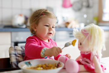 Adorable baby girl eating from fork vegetables and pasta. Little child feeding and playing with toy...