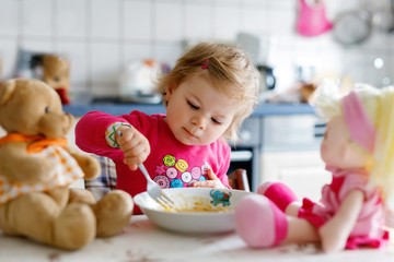 Adorable baby girl eating from fork vegetables and pasta. Little child feeding and playing with toy doll