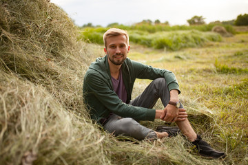 Naklejka na ściany i meble Young smiling man sits near haystack on meadow.