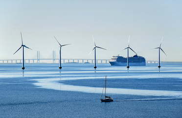 Very large passenger ship and a small sailboat pass offshore wind turbines near the Oresund Bridge...